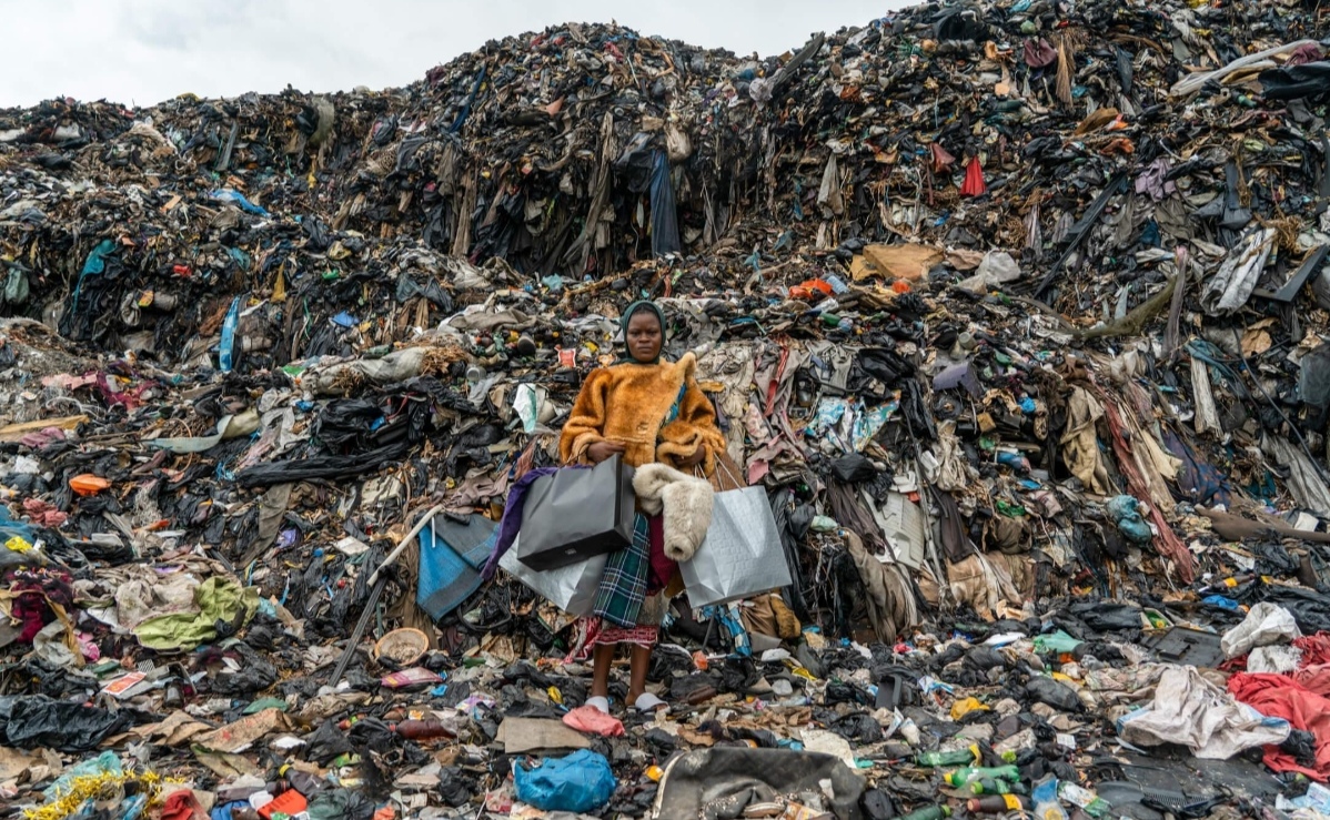 A person standing in front of a massive textile waste landfill while holding shopping bags, illustrating the environmental impact of fast fashion and clothing overconsumption.
